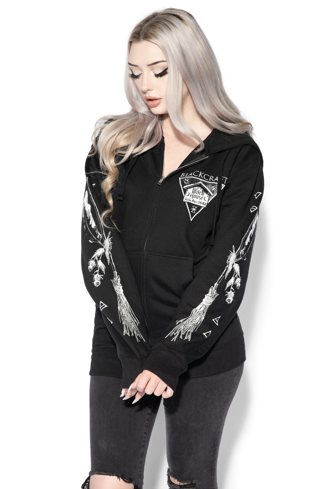 A blond model wearing a classic black zip up hoodie with Salem Massachusetts artwork along the front, back, and along the sleeves. 
