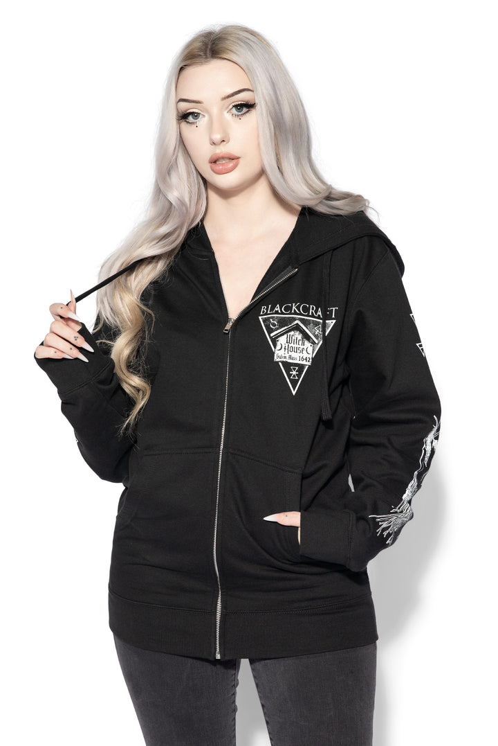 A blond model wearing a classic black zip up hoodie with Salem Massachusetts artwork along the front, back, and along the sleeves. 