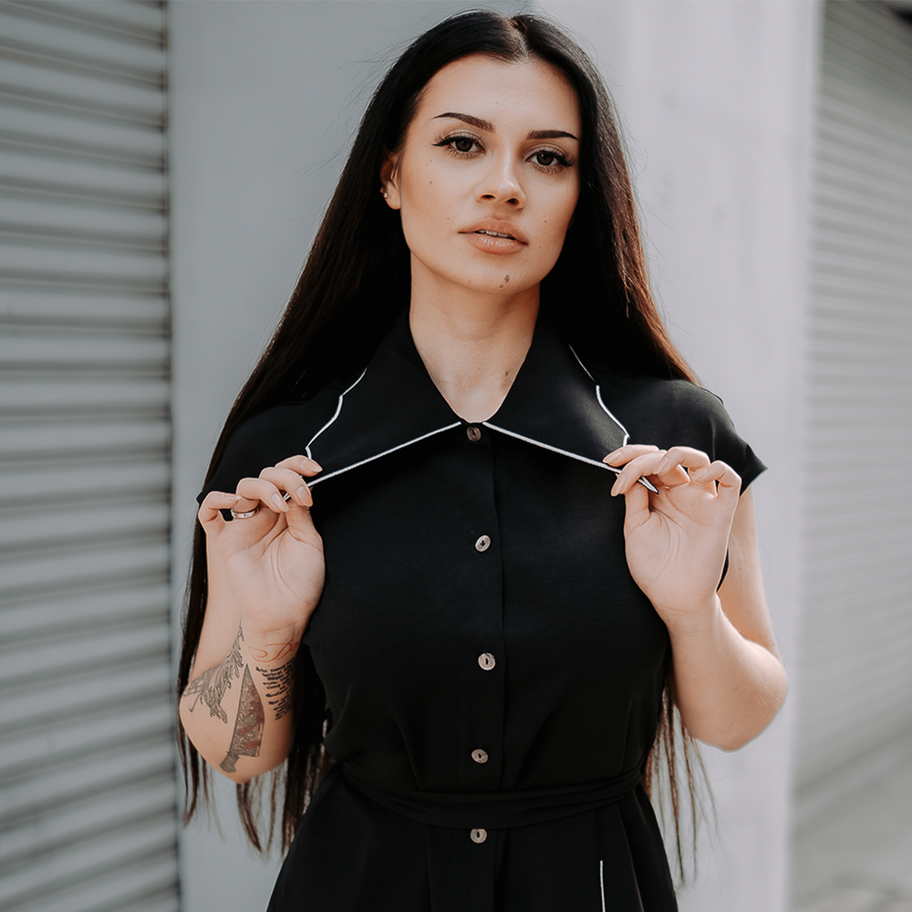 A model wearing a classic black shirt dress with a spider web inspired collar with contrast white trim, an embroidered spider detail, a button front, short sleeves, and an adjustable waist tie. 