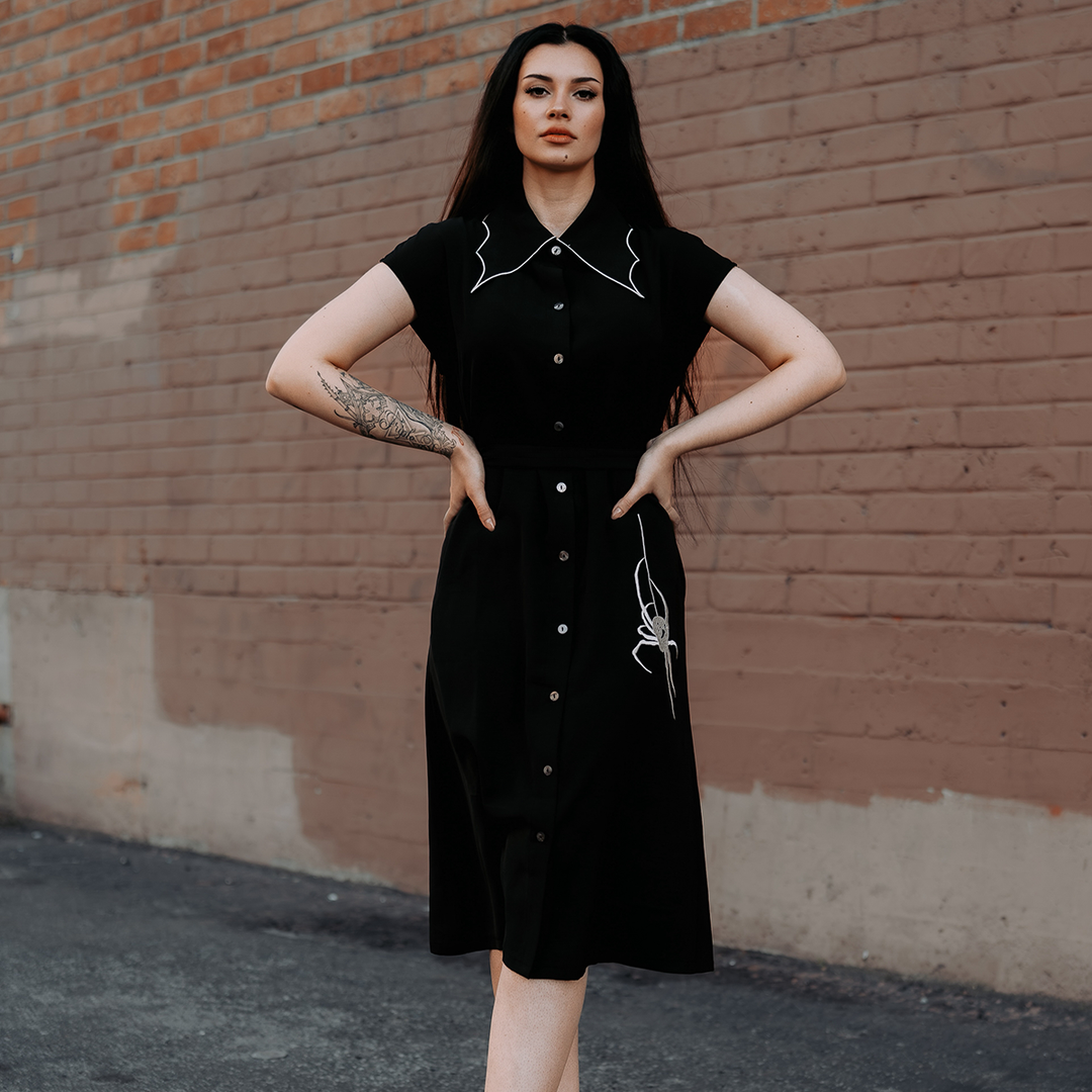 A model wearing a classic black shirt dress with a spider web inspired collar with contrast white trim, an embroidered spider detail, a button front, short sleeves, and an adjustable waist tie. 