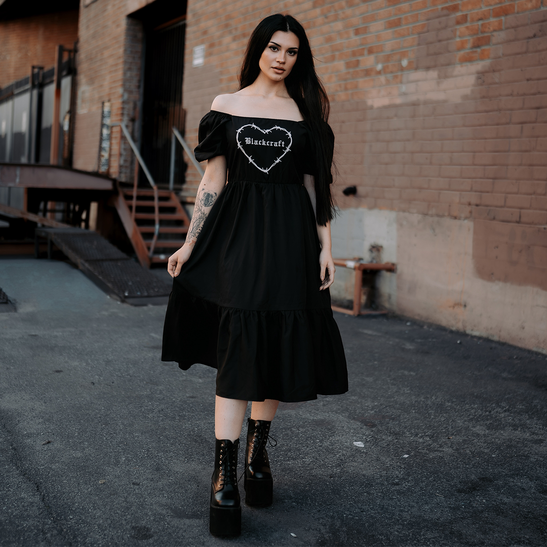 A model standing outside wearing a black gothic maxi dress with short puff sleeves, a long flowy tiered skirt, and a half smocked bodice at the back for a slim fit.