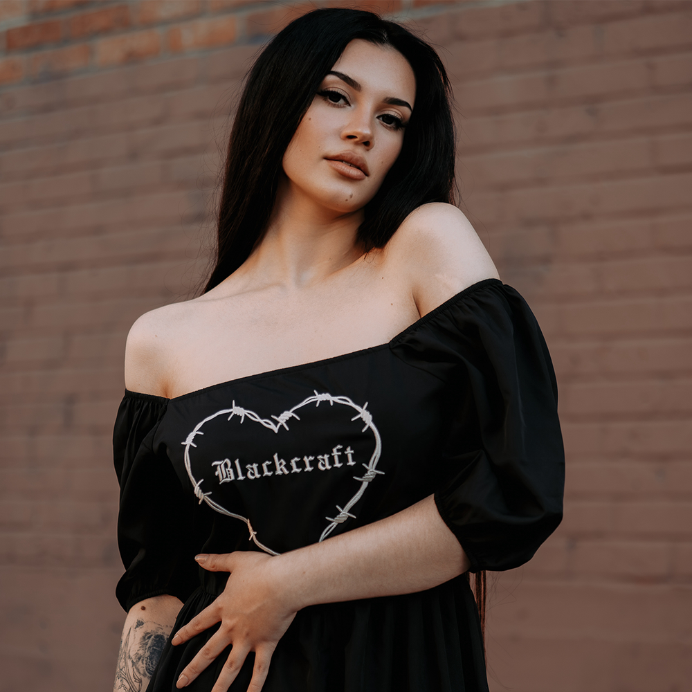 A model standing outside wearing a black gothic maxi dress with short puff sleeves, a long flowy tiered skirt, and a half smocked bodice at the back for a slim fit.