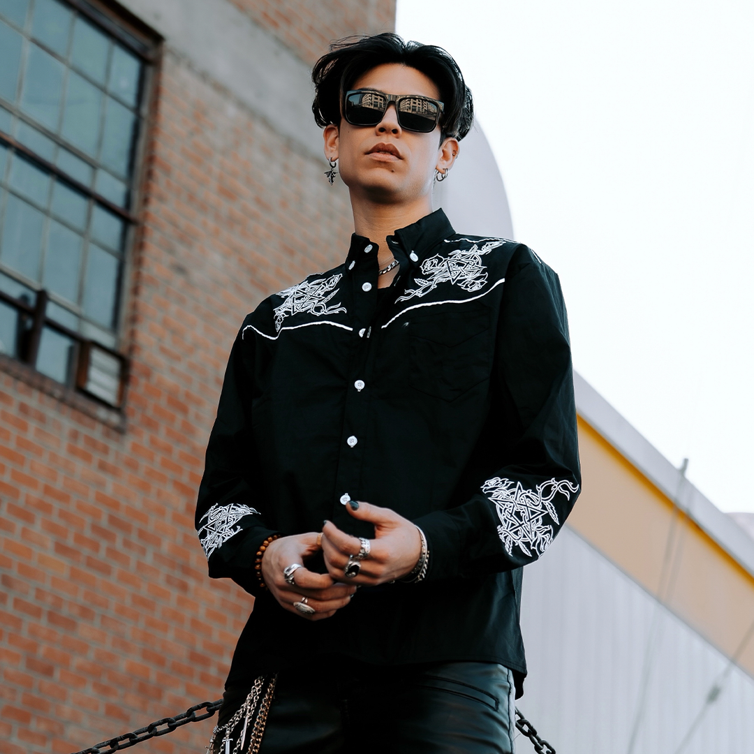 A male model wearing a black cotton western style button front shirt with a collared neckline and western style embroidered details in white, from Blackcraft. 