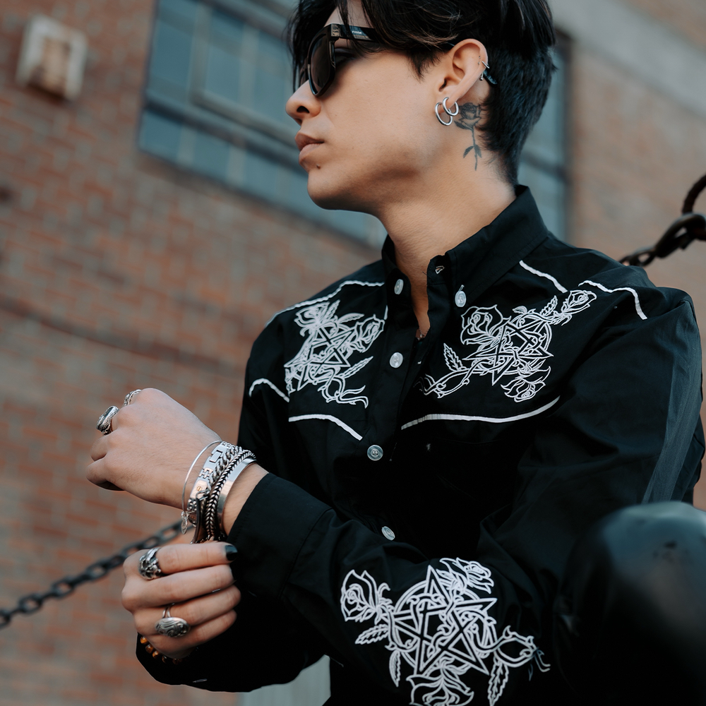 A male model wearing a black cotton western style button front shirt with a collared neckline and western style embroidered details in white, from Blackcraft. 