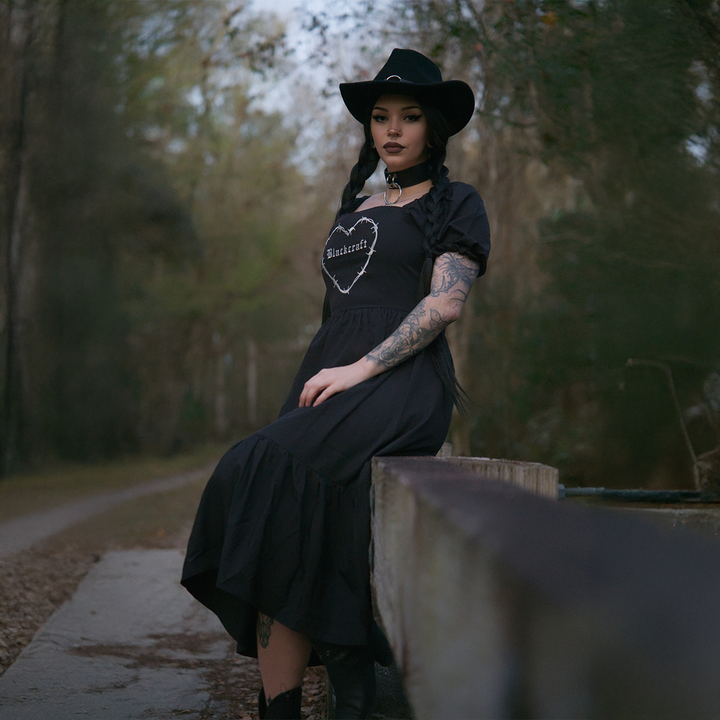 A model standing outside wearing a black gothic maxi dress with short puff sleeves, a long flowy tiered skirt, and a half smocked bodice at the back for a slim fit.