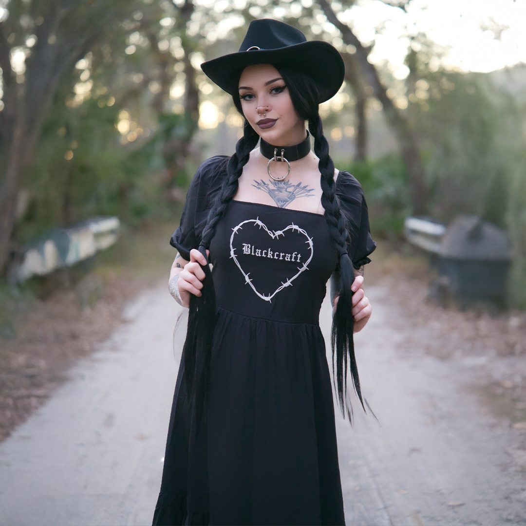 A model standing outside wearing a black gothic maxi dress with short puff sleeves, a long flowy tiered skirt, and a half smocked bodice at the back for a slim fit.