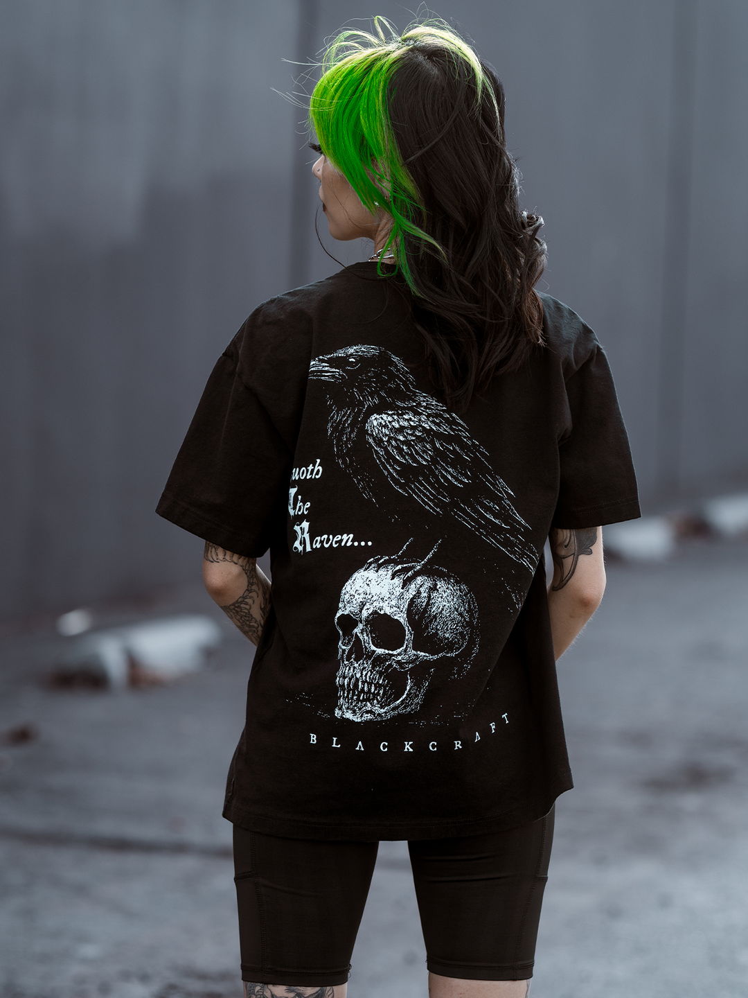 A green haired model standing outside wearing a classic black cotton boxy tee with Edgar Allen poe and the Raven artwork, from Blackcraft.
