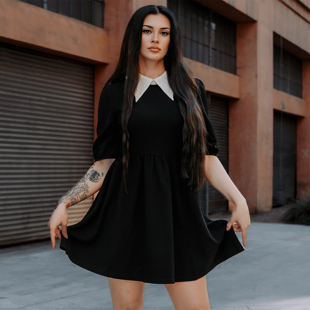 A dark haired woman standing outside wearing a black puff sleeve mini dress with contrast white pointed collar. 