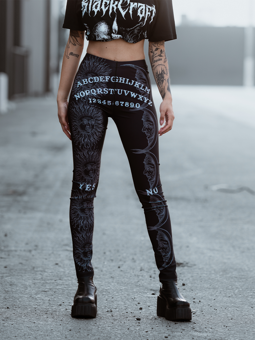 A model standing outside wearing classic women's black leggings with an allover ouija print. 