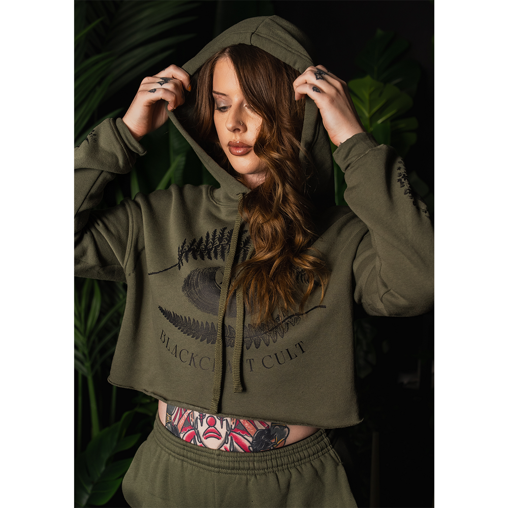 A model wearing a military green Blackcraft cropped hoodie with custom plant witch artwork on the front and along the sleeves.