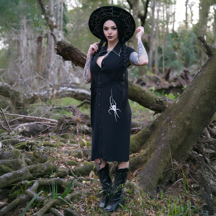 A model wearing a classic black shirt dress with a spider web inspired collar with contrast white trim, an embroidered spider detail, a button front, short sleeves, and an adjustable waist tie. 