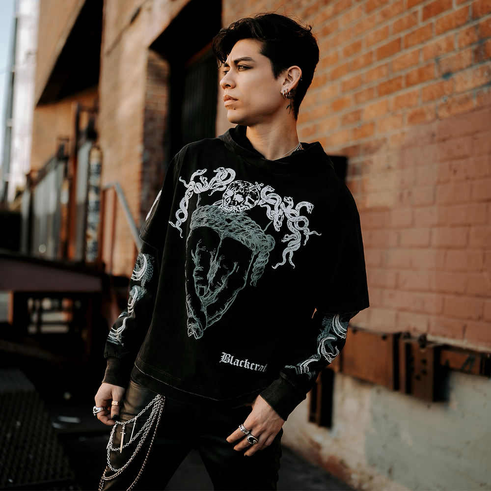 A model standing outside wearing a washed black long sleeve twofer pullover hoodie with custom Blackcraft artwork on the front and along the sleeves.