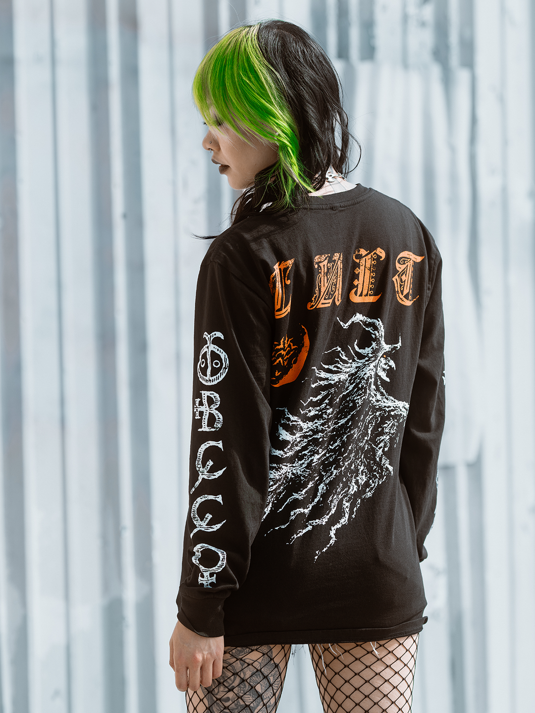 A green haired model standing outside wearing a classic black cotton long sleeve tee with orange and white witch artwork, from Blackcraft.