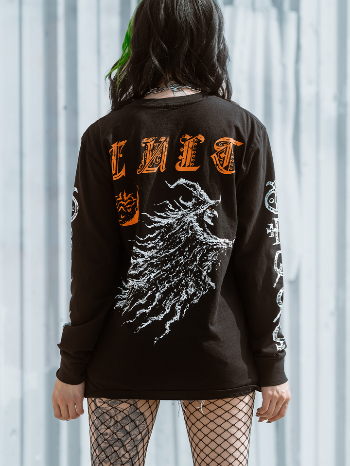 A green haired model standing outside wearing a classic black cotton long sleeve tee with orange and white witch artwork, from Blackcraft.