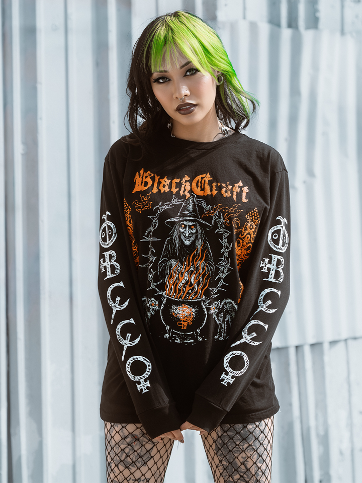 A green haired model standing outside wearing a classic black cotton long sleeve tee with orange and white witch artwork, from Blackcraft.