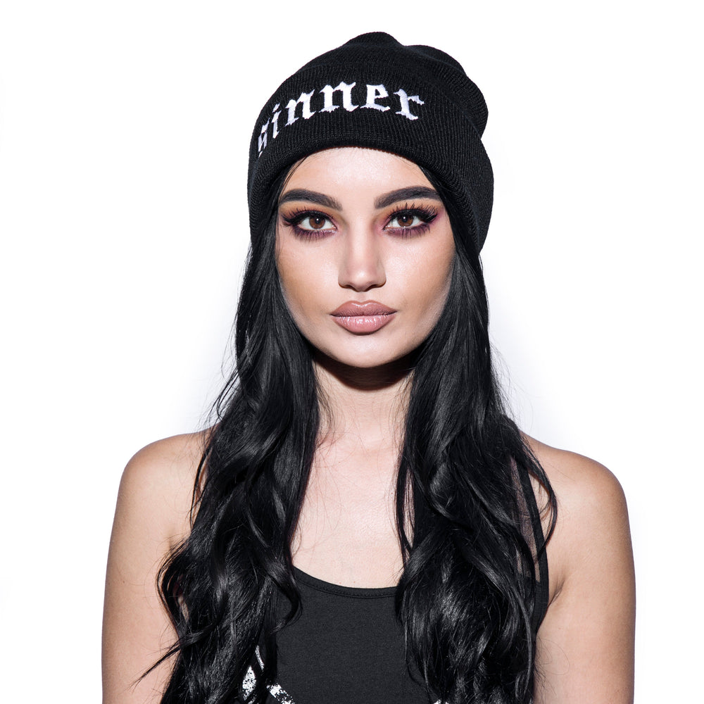 Model wearing a black beanie with sinner embroidery on front flap.
