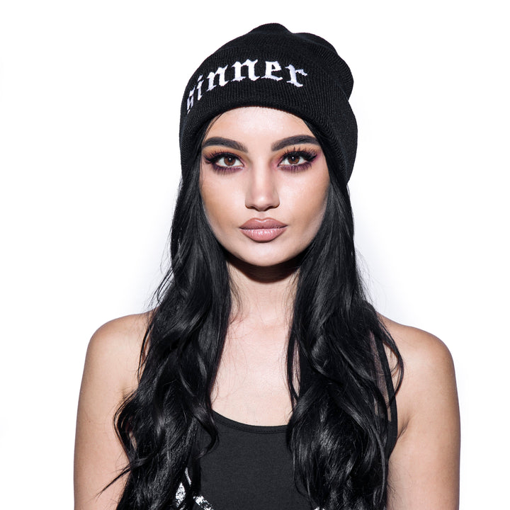 Model wearing a black beanie with sinner embroidery on front flap.