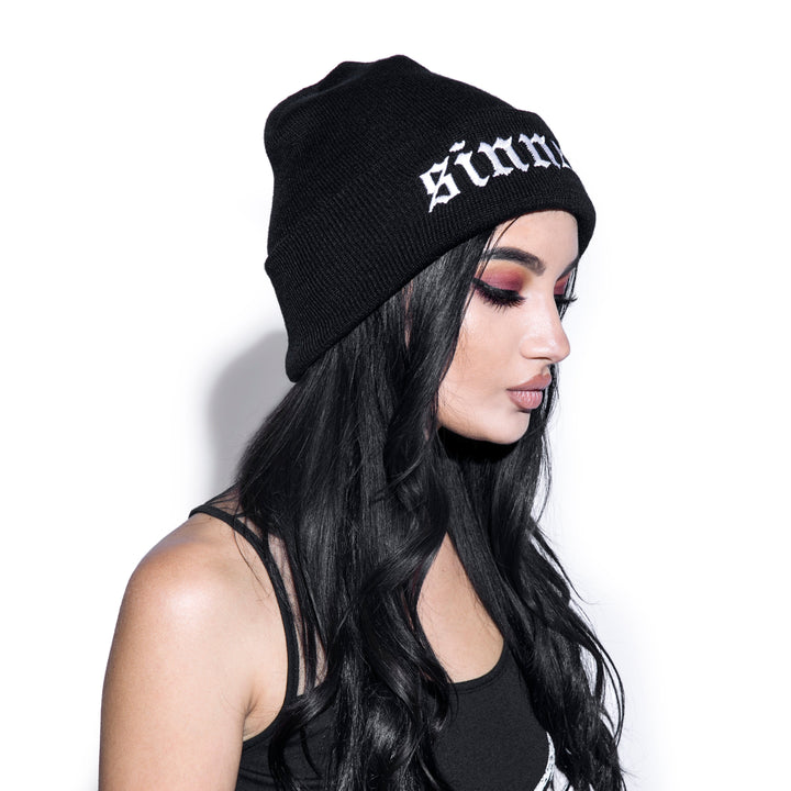 Model wearing a black beanie with sinner embroidery on front flap.