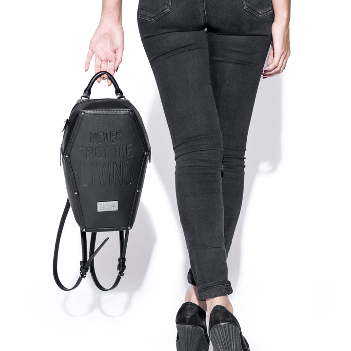 Black faux leather coffin shaped backpack with Never Trust the Living embossed on front. 