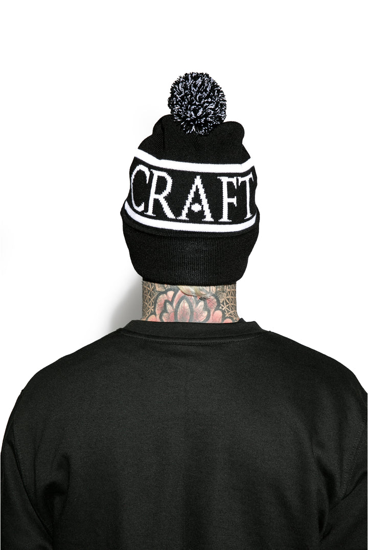 Model wearing a black bobble beanie with Blackcraft across the circumference.