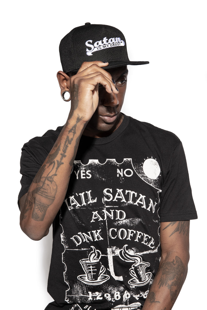 A model wearing a black snapback hat with white embroidered Satan is my Daddy on the front. 